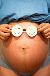 Midsection of pregnant woman in nightwear with smiley face eyeglasses standing against wall