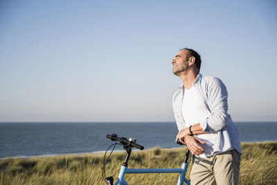 Man with eyes closed leaning on bicycle at beach against clear sky