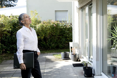 Senior woman holding laptop standing with hand in pocket looking at house