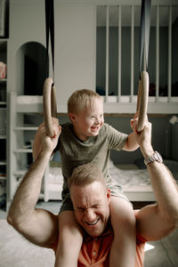 Man carrying son with down syndrome on shoulders while holding gymnastic rings in bedroom