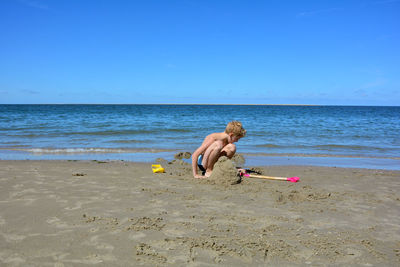 A blond boy with swimming trunks builds a sand castle in the sand on the beach in front of the sea