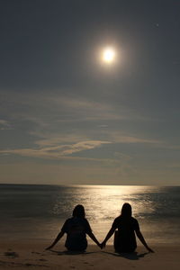 Rear view of silhouette friends sitting on shore at beach against sky during sunset