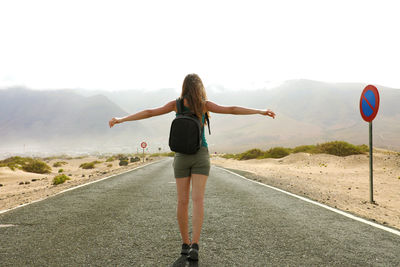 Rear view of woman with arms outstretched walking on road against sky