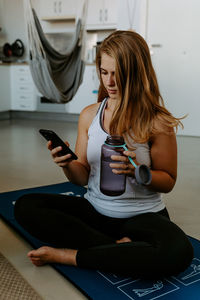 Smiling young female in sportswear holding water bottle and taking selfie on modern mobile phone during home workout