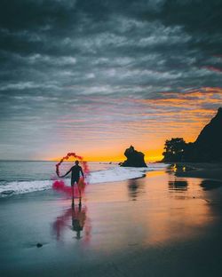 Full length of man holding smoke bomb at beach against cloudy sky during sunset