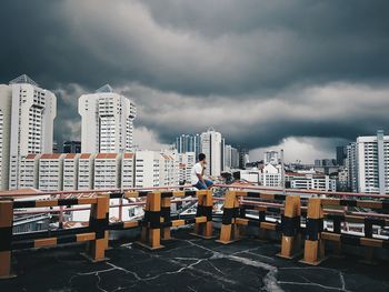 Man sitting on railing of building terrace while looking at buildings against cloudy sky