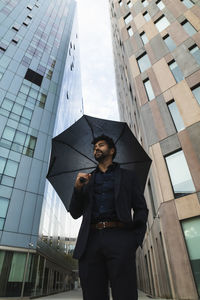 Entrepreneur with hand in pocket holding umbrella against building in city