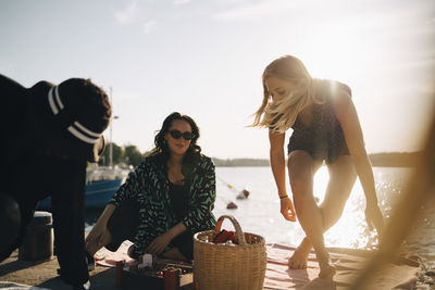 Friends with picnic basket sitting at jetty against lake during summer