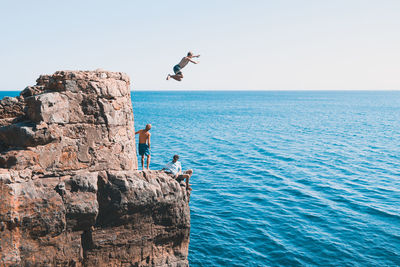 People jumping on rock by sea against clear sky