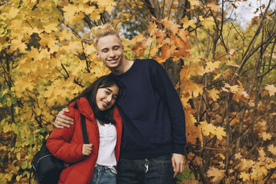 Smiling teenage friends standing against maple trees during autumn