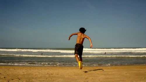 Shirtless boy running towards sea at beach against clear sky