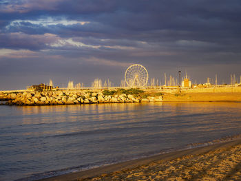 Ferris wheel by sea against cloudy sky