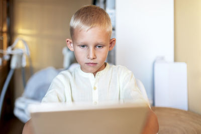 Side view of concentrated blond haired little boy in white shirt browsing internet on tablet in bright room in sunlight