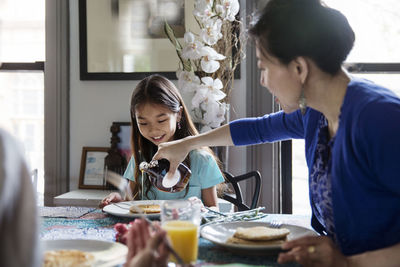 Mother pouring maple syrup on pancakes for daughter during breakfast