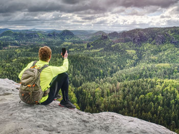 Trail runner takes rest, makes photos by his smart phone. view into spring forest below low mountain