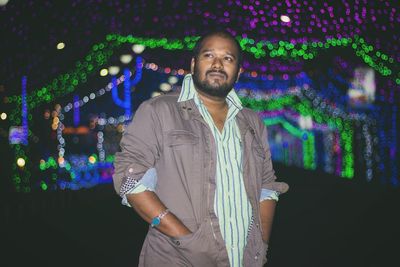 Thoughtful man with hands in pockets standing against illuminated lights