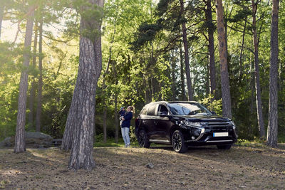 Young woman carrying baby boy while standing by car amidst trees