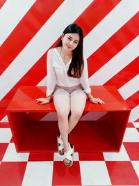 Portrait of young woman sitting on red seat against striped wall