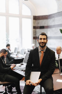 Portrait of smiling male lawyer holding laptop sitting on desk in office