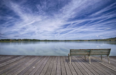 Empty bench on pier by lake against sky