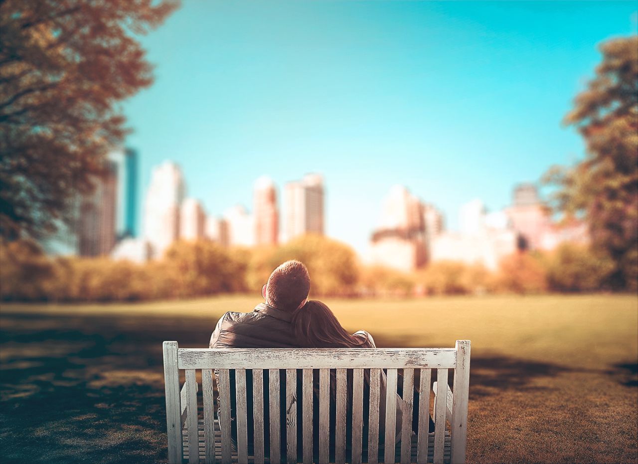 REAR VIEW OF MAN SITTING ON SEAT AT PARK AGAINST SKY