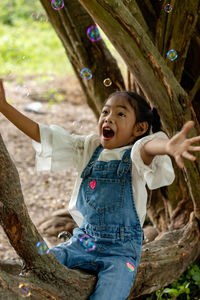 Happy girl playing with bubble while sitting on tree trunk