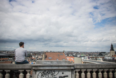 Rear view of man sitting by buildings against sky