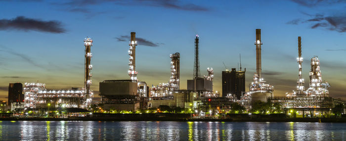 Illuminated factory by river against sky