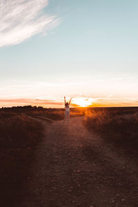 Woman with arms raised standing on road against sky during sunset