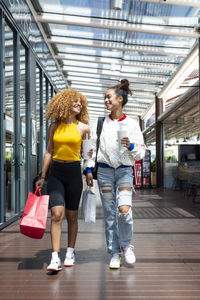 Positive african american female friends with shopping paper bags walking along mall and looking at each other