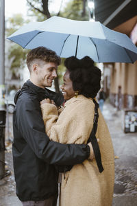 Side view of happy multiracial couple standing under umbrella at street