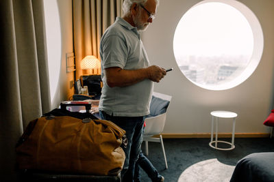 Senior man using smart phone while standing against window in hotel room