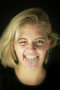 Close-up portrait of young woman sticking out tongue against black background