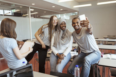 Smiling man taking selfie with female friends on smart phone at classroom