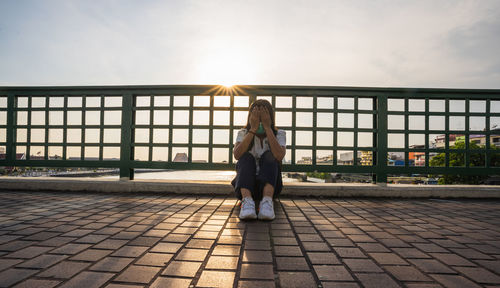 Asian woman wearing surgical mask sits on bridge, hands holding her head due to unemployment stress.