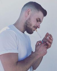Side view of young man lighting cigarette with lighter against wall