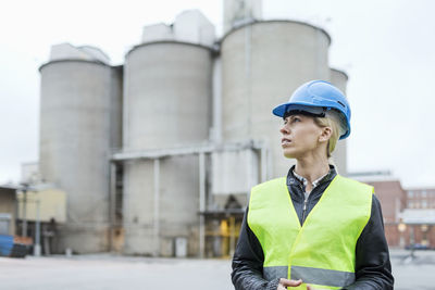 Female manual worker looking away while standing against cement silos
