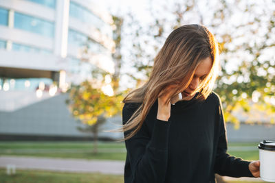 Portrait of young woman student with long hair using mobile phone in city park in golden hour