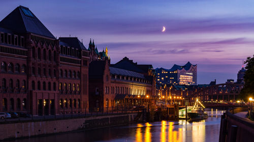 Illuminated city by river against sky at dusk