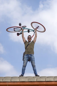 Low angle view of mid adult man carrying bicycle while standing against sky
