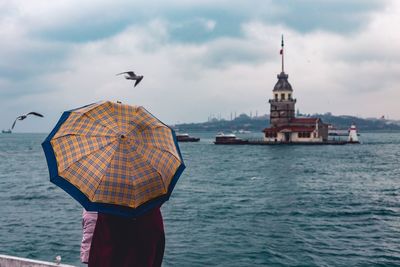 Rear view of person with umbrella standing by sea against cloudy sky