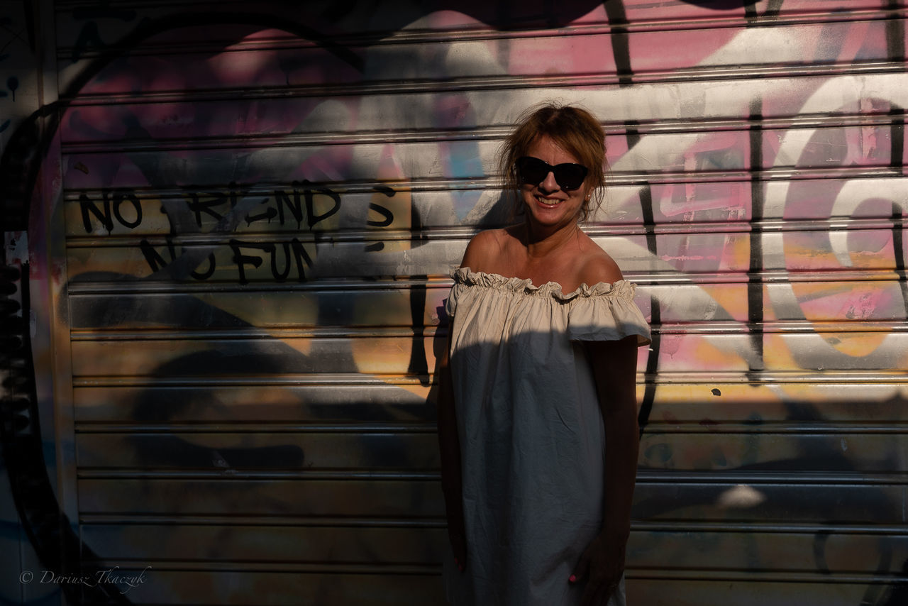 sunglasses, glasses, one person, fashion, adult, young adult, standing, front view, women, graffiti, architecture, lifestyles, casual clothing, portrait, clothing, three quarter length, looking at camera, leisure activity, day, city, outdoors, smiling, hairstyle, built structure