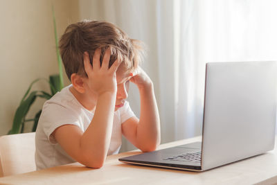 Desperate child sits at desk, looks sadly at computer and holds his head with hands. online learning