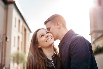 Man kissing woman on cheek while standing amidst buildings against sky