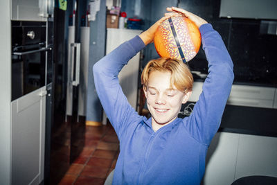 Smiling boy holding sports ball on head in kitchen