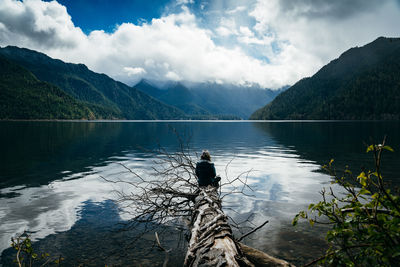 Woman sitting on broken tree while looking at lake and mountains against sky