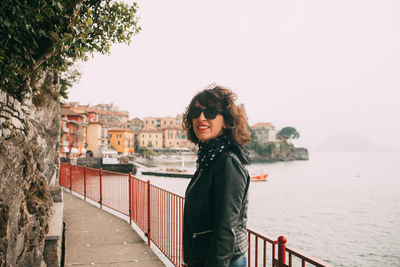 Portrait of smiling mid adult woman standing on footpath by sea against clear sky