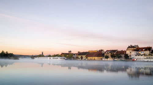 Buildings by river against sky at sunset