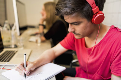 Young businessman wearing headphones while writing on book with colleague in background at creative office