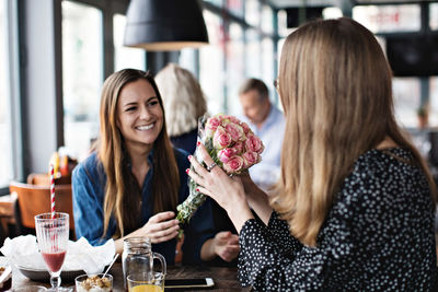 Smiling young woman giving fresh flower bouquet to female friend sitting at restaurant